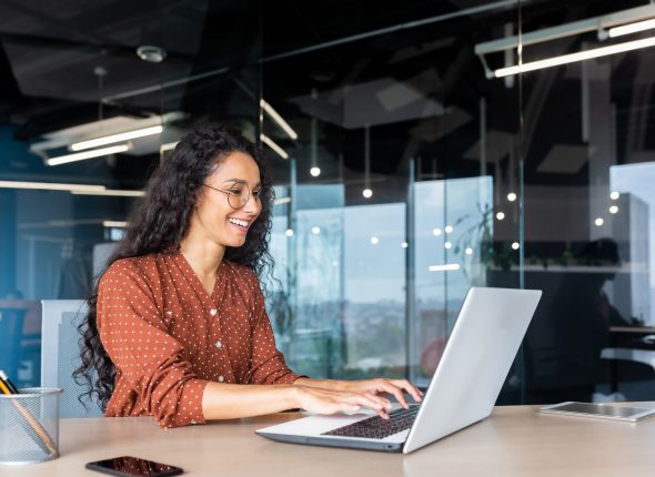 Happy and smiling hispanic businesswoman typing on laptop, office worker with curly hair and glasses happy with achievement results, at work inside office building